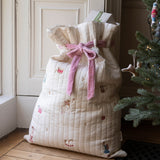 A quilted cream Christmas stocking filled with gifts, tied with a pink ribbon, standing on a wooden floor next to a decorated Christmas tree.