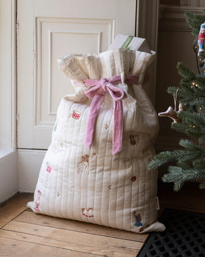 A quilted cream Christmas stocking filled with gifts, tied with a pink ribbon, standing on a wooden floor next to a decorated Christmas tree.