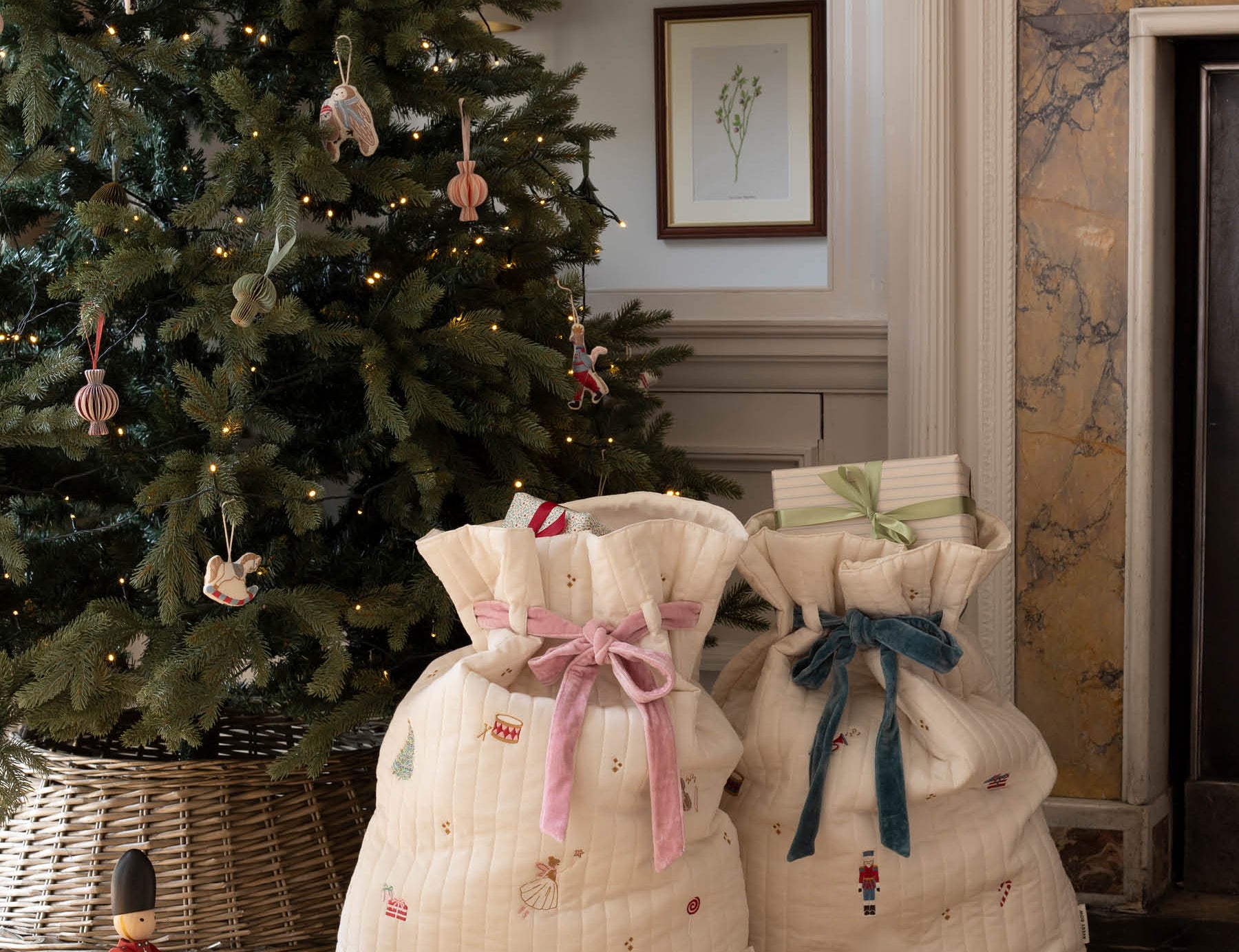 Two quilted cream fabric gift bags with festive prints and ribbon bows, placed on a woven rug in front of a decorated Christmas tree and fireplace.