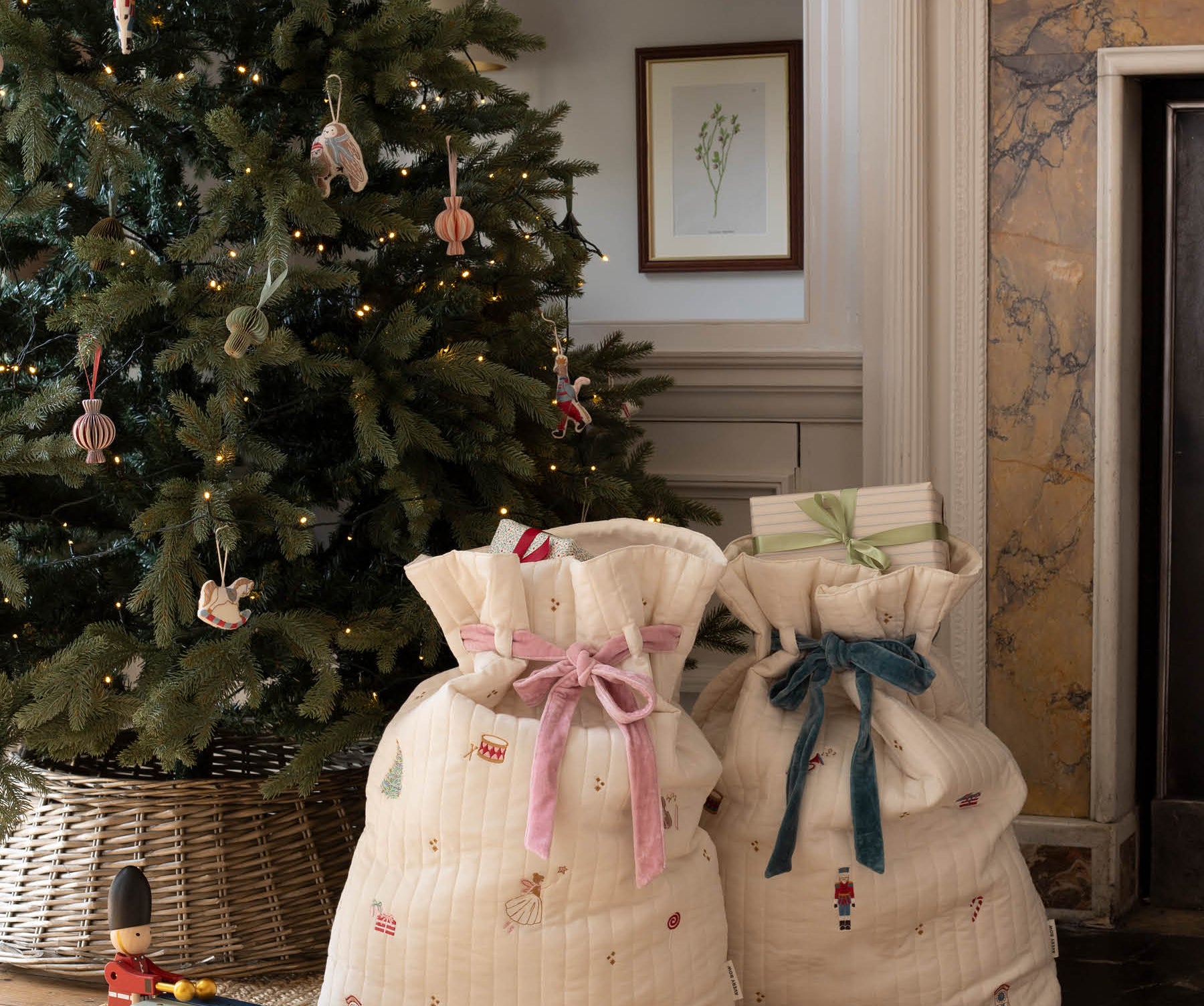 Two quilted cream fabric gift bags with festive prints and ribbon bows, placed on a woven rug in front of a decorated Christmas tree and fireplace.