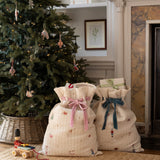Two quilted cream fabric gift bags with festive prints and ribbon bows, placed on a woven rug in front of a decorated Christmas tree and fireplace.