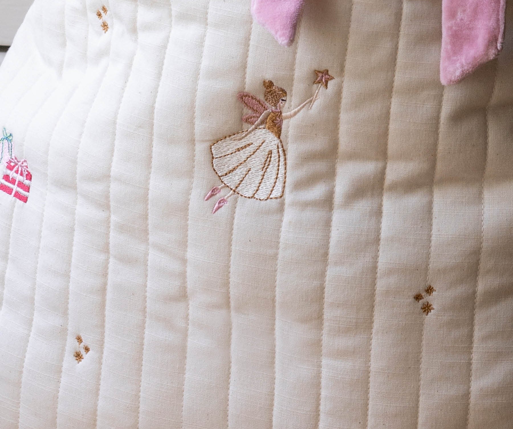 Close-up of a quilted cream fabric item with embroidered fairy and rocking horse motifs, plus pink ribbon ties at the top.
