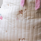 Close-up of a quilted cream fabric item with embroidered fairy and rocking horse motifs, plus pink ribbon ties at the top.