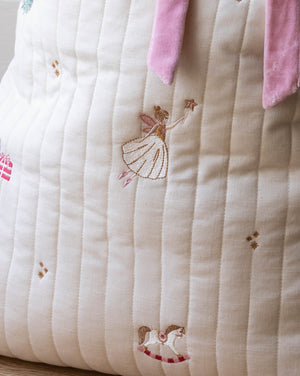 Close-up of a quilted cream fabric item with embroidered fairy and rocking horse motifs, plus pink ribbon ties at the top.