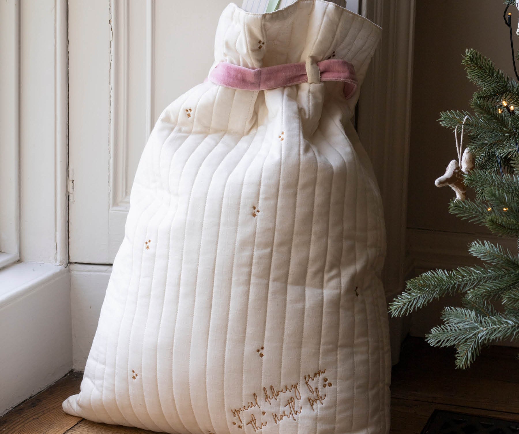 A large quilted cream gift bag with a pink ribbon tied around the neck, placed on a wooden floor next to a Christmas tree.