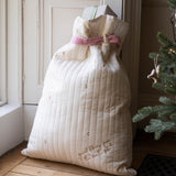 A large quilted cream gift bag with a pink ribbon tied around the neck, placed on a wooden floor next to a Christmas tree.