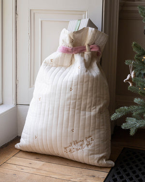 A large quilted cream gift bag with a pink ribbon tied around the neck, placed on a wooden floor next to a Christmas tree.