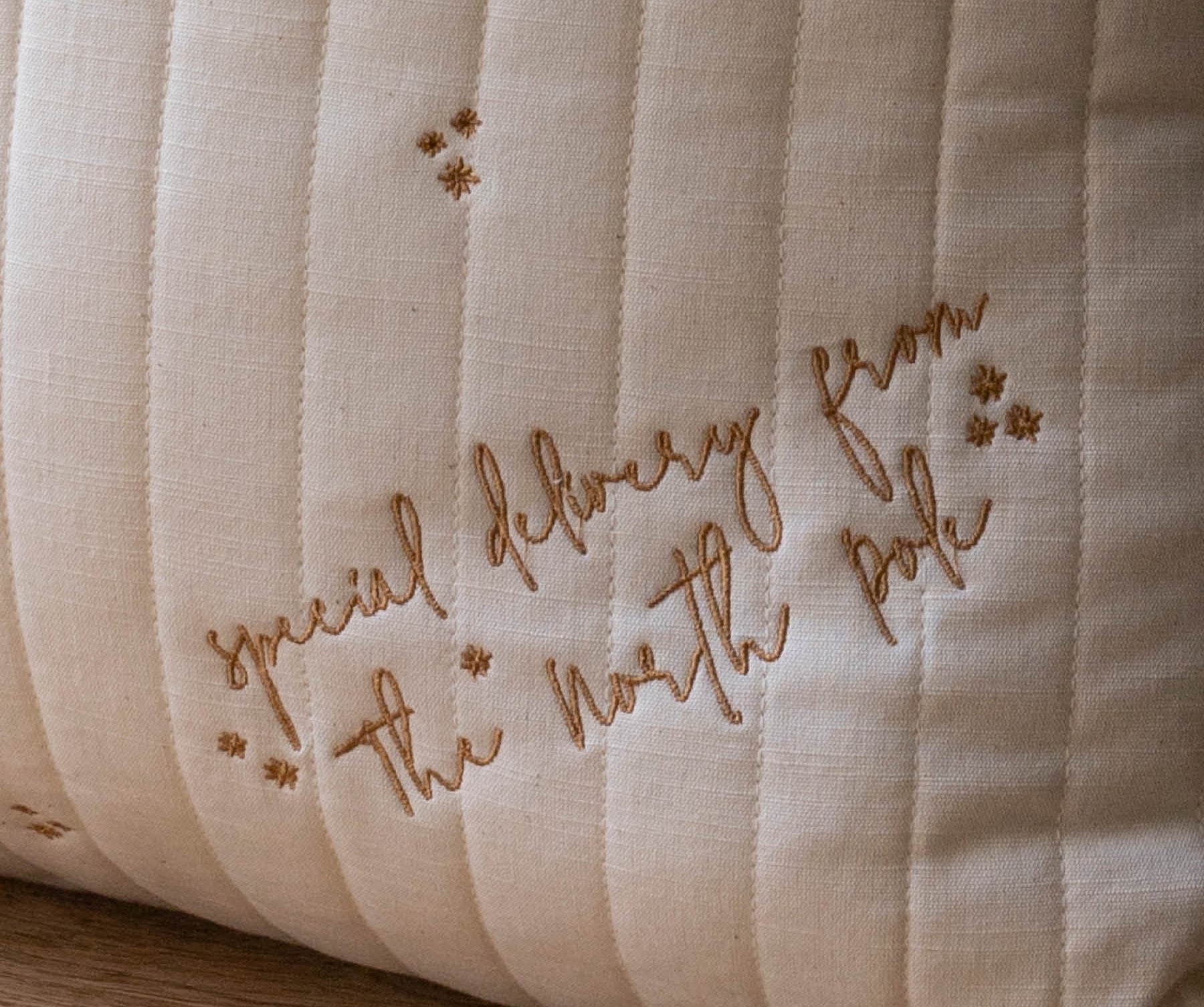 A close-up of a cream-colored quilted cushion with vertical channel stitching and embroidered brown script reading 'special delivery from the north pole' alongside small star motifs, resting on a wooden surface.