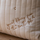 A close-up of a cream-colored quilted cushion with vertical channel stitching and embroidered brown script reading 'special delivery from the north pole' alongside small star motifs, resting on a wooden surface.