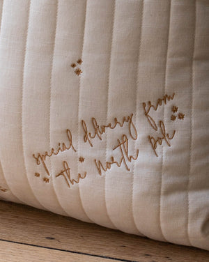 A close-up of a cream-colored quilted cushion with vertical channel stitching and embroidered brown script reading 'special delivery from the north pole' alongside small star motifs, resting on a wooden surface.
