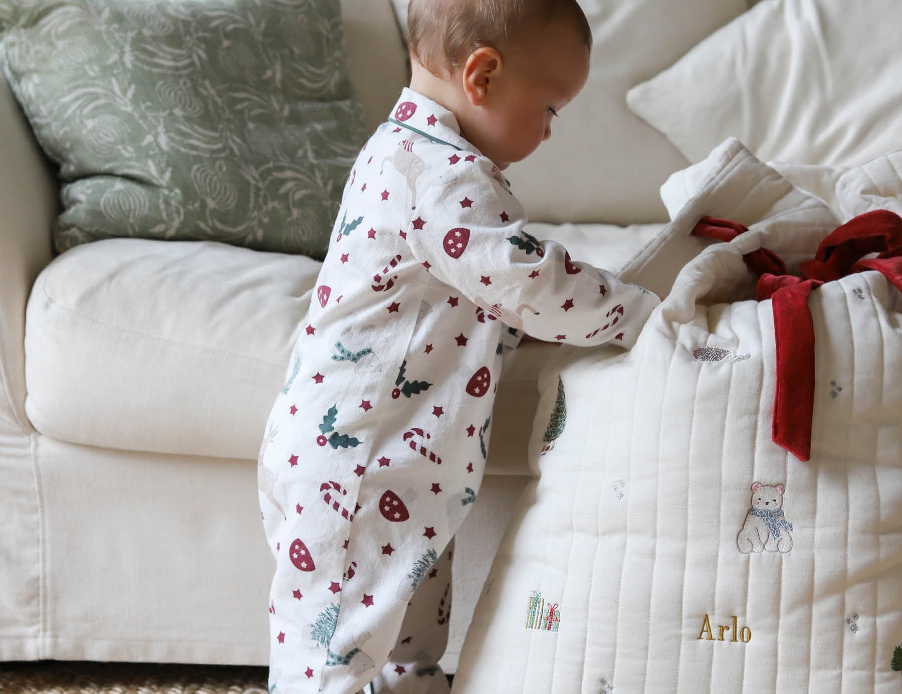 A baby wearing a white footless pajama romper with a red and green Christmas print stands next to a quilted white baby blanket with personalized gold text and a red ribbon, in a cozy living room setting.