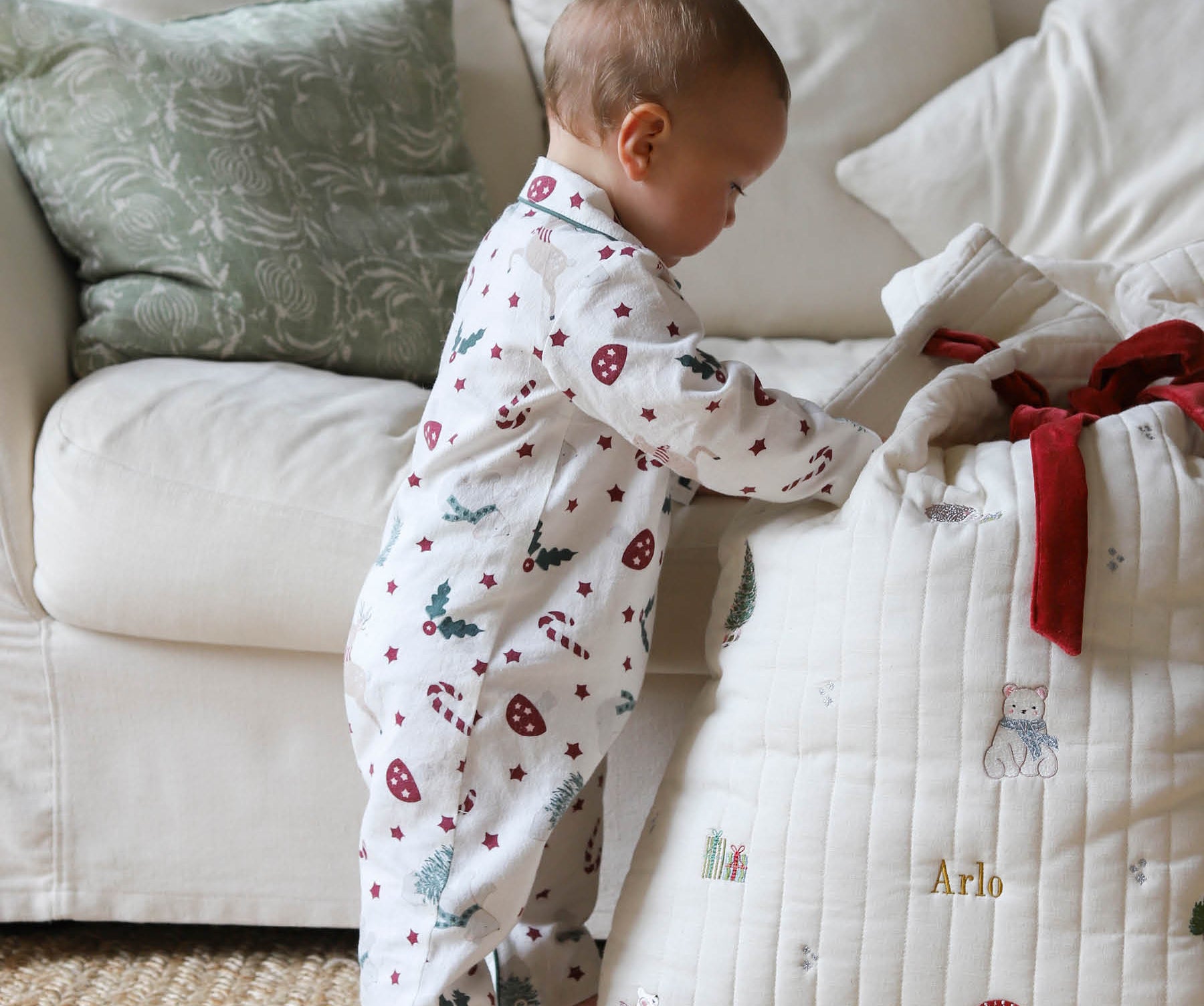 A baby wearing a white footless pajama romper with a red and green Christmas print stands next to a quilted white baby blanket with personalized gold text and a red ribbon, in a cozy living room setting.