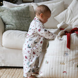 A baby wearing a white footless pajama romper with a red and green Christmas print stands next to a quilted white baby blanket with personalized gold text and a red ribbon, in a cozy living room setting.