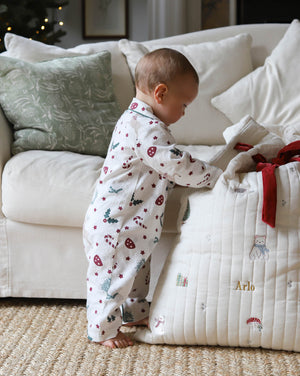 A baby wearing a white footless pajama romper with a red and green Christmas print stands next to a quilted white baby blanket with personalized gold text and a red ribbon, in a cozy living room setting.