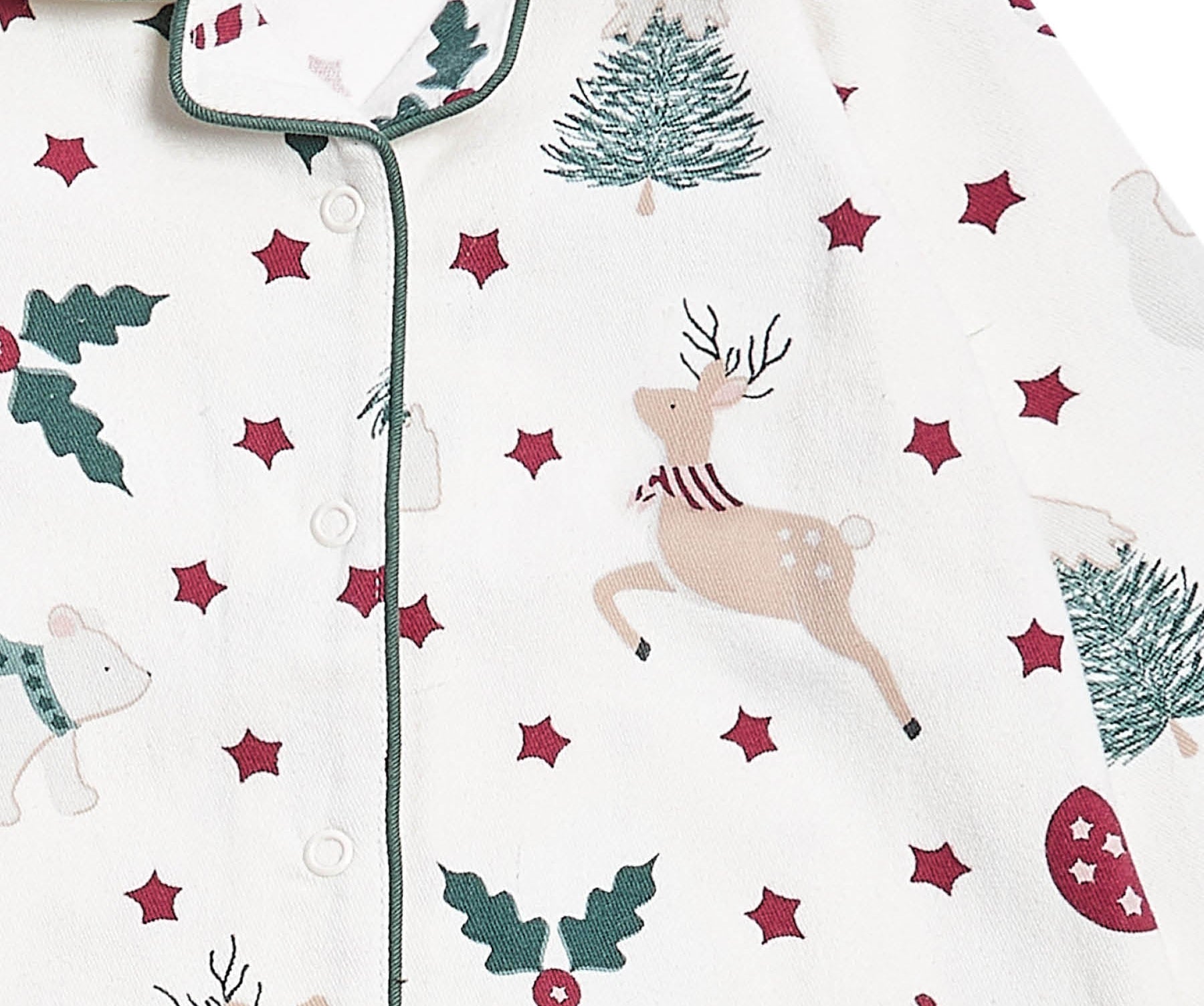 A close-up of a baby pajama top by Avery Row, featuring a festive Christmas print with a beige deer, green trees and holly, and red stars on an off-white background.