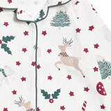 A close-up of a baby pajama top by Avery Row, featuring a festive Christmas print with a beige deer, green trees and holly, and red stars on an off-white background.