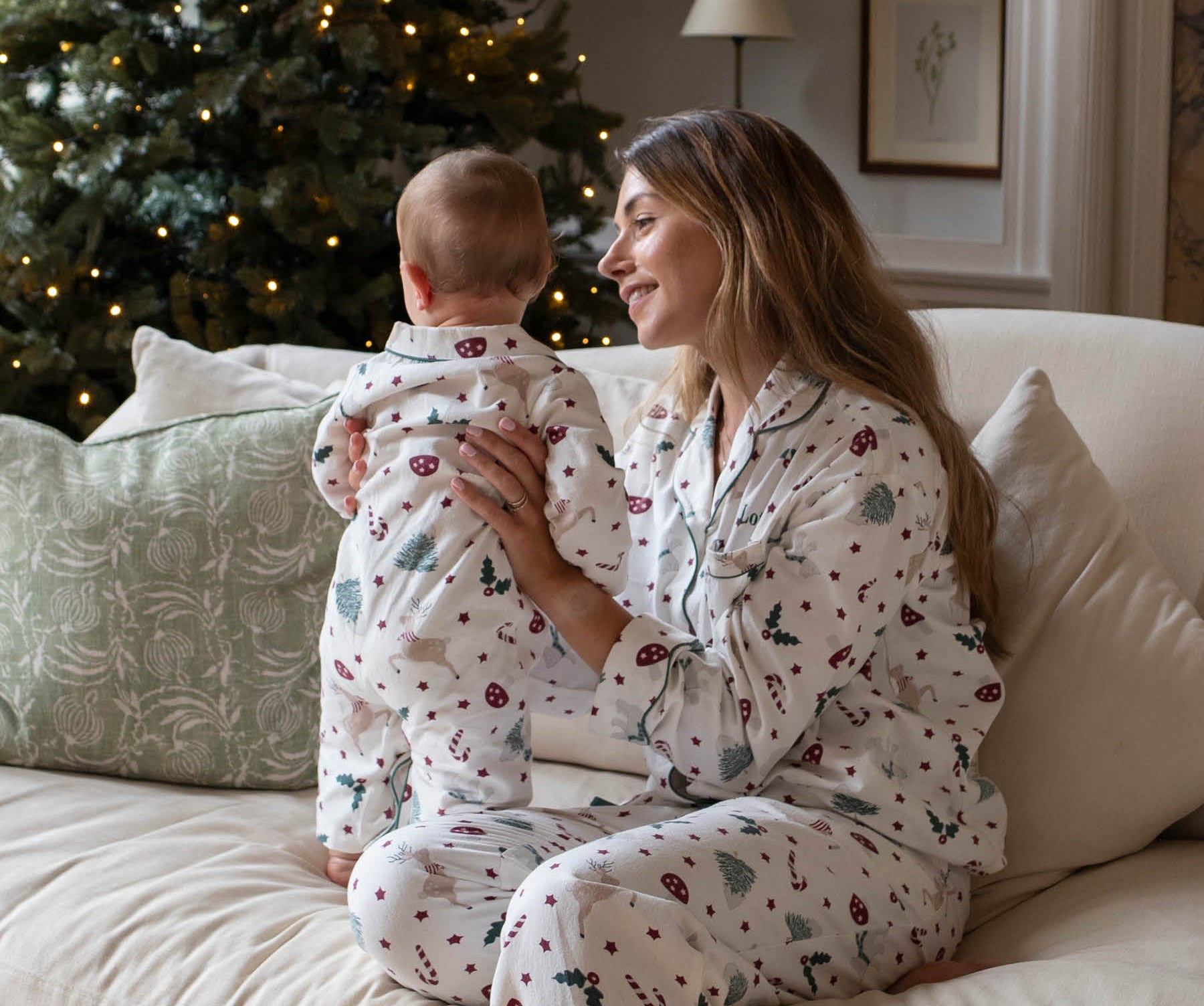 A woman and a baby sitting on a beige couch, both wearing matching white Christmas-themed pajamas with a red and green holiday print. A decorated Christmas tree with warm lights is visible in the background.