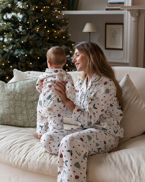 A woman and a baby sitting on a beige couch, both wearing matching white Christmas-themed pajamas with a red and green holiday print. A decorated Christmas tree with warm lights is visible in the background.