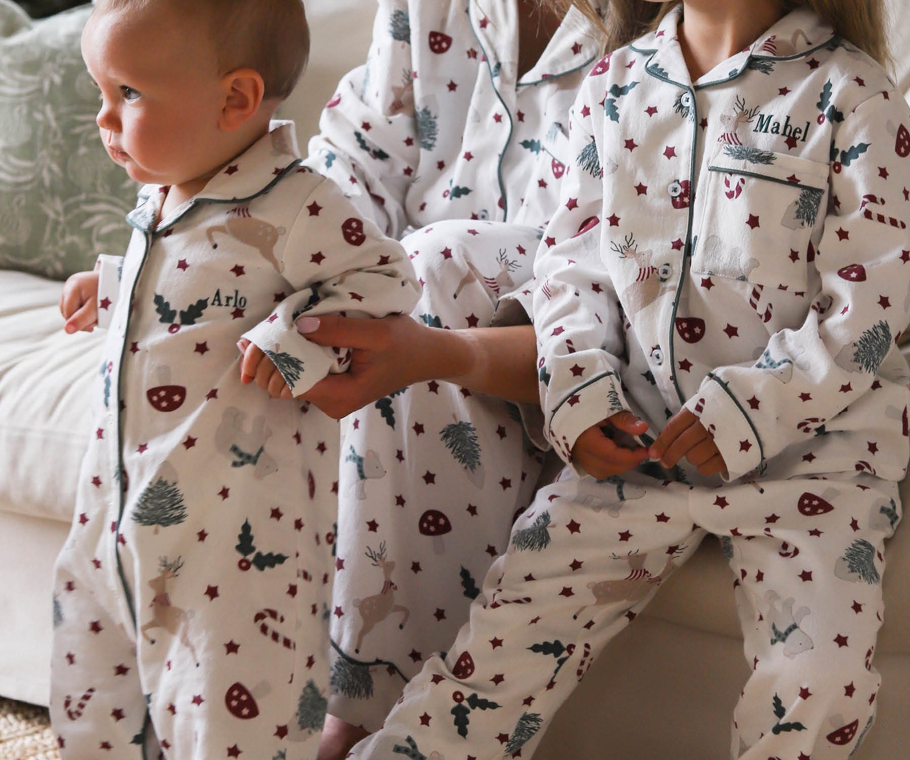 Three children wearing matching white holiday pajamas with a festive print of candy canes, Christmas trees, and stars, sitting on a couch in a cozy living room setting.