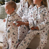 Three children wearing matching white holiday pajamas with a festive print of candy canes, Christmas trees, and stars, sitting on a couch in a cozy living room setting.