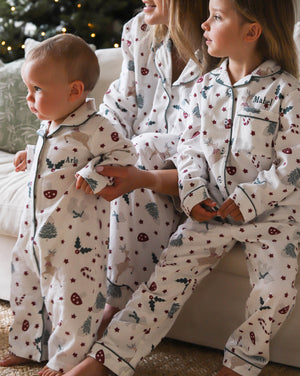 Three children wearing matching white holiday pajamas with a festive print of candy canes, Christmas trees, and stars, sitting on a couch in a cozy living room setting.
