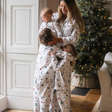 A woman and two children wearing matching Christmas-themed pajamas with a festive print, standing in a cozy room next to a decorated Christmas tree.