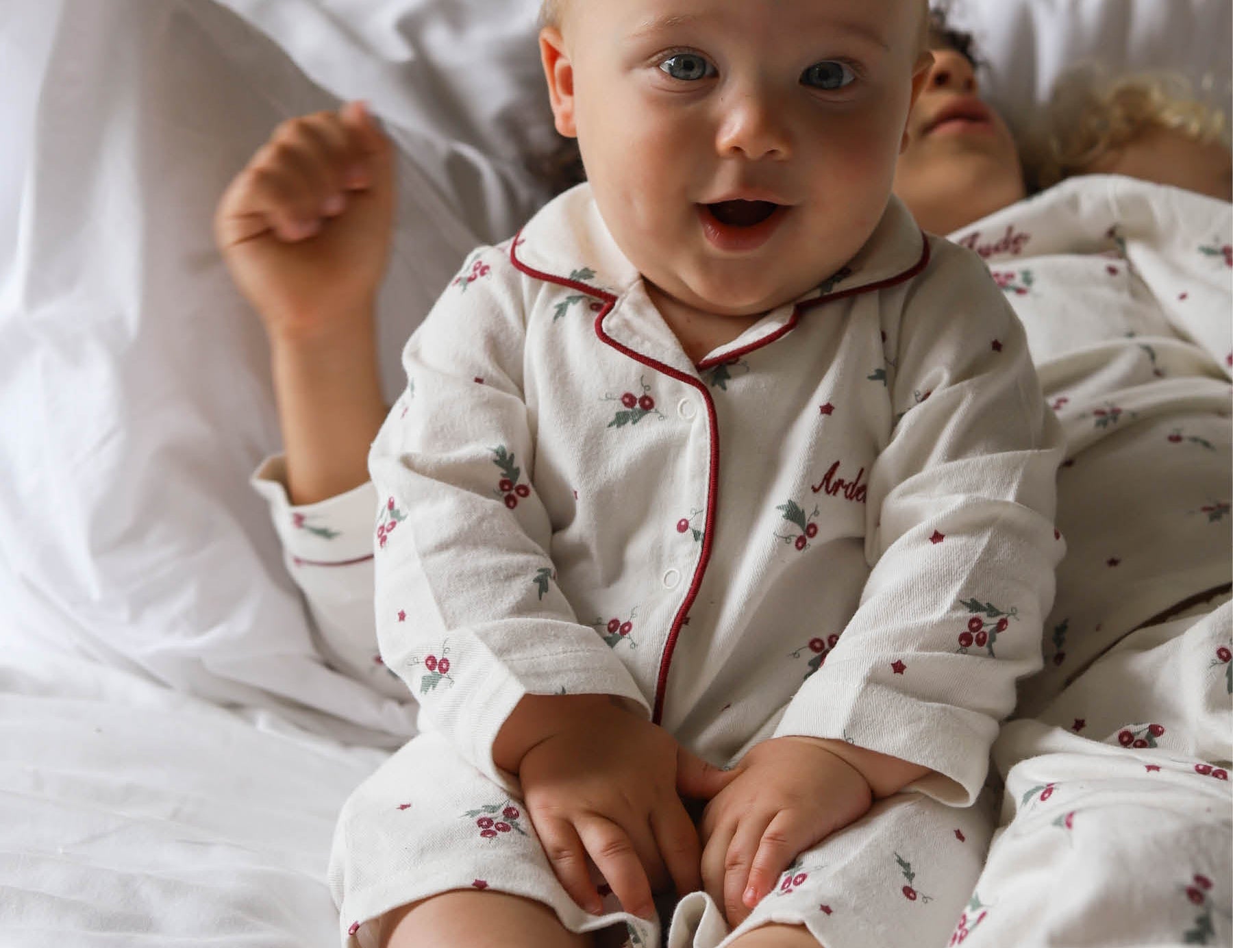 A baby sitting up in bed wearing a white long-sleeve pajama set with red piping and a small cherry print. The baby is smiling and looking directly at the camera, with white pillows and bedding in the background.