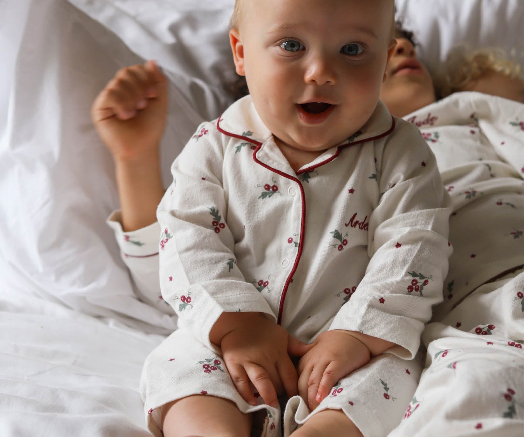 A baby sitting up in bed wearing a white long-sleeve pajama set with red piping and a small cherry print. The baby is smiling and looking directly at the camera, with white pillows and bedding in the background.