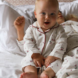 A baby sitting up in bed wearing a white long-sleeve pajama set with red piping and a small cherry print. The baby is smiling and looking directly at the camera, with white pillows and bedding in the background.