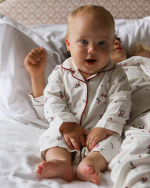 A baby sitting up in bed wearing a white long-sleeve pajama set with red piping and a small cherry print. The baby is smiling and looking directly at the camera, with white pillows and bedding in the background.