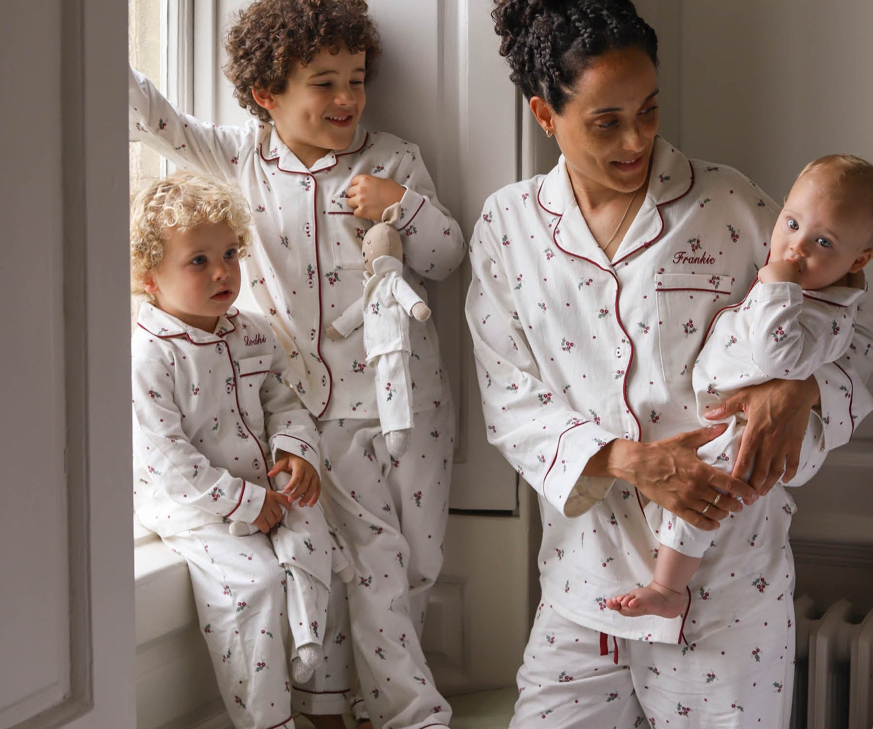 A family of four wearing matching white patterned pajamas with red piping, posed in a bright bedroom setting. The set includes an adult and three children, all wearing coordinated long-sleeve tops and pants with red trim and a small repeating motif.
