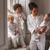A family of four wearing matching white patterned pajamas with red piping, posed in a bright bedroom setting. The set includes an adult and three children, all wearing coordinated long-sleeve tops and pants with red trim and a small repeating motif.
