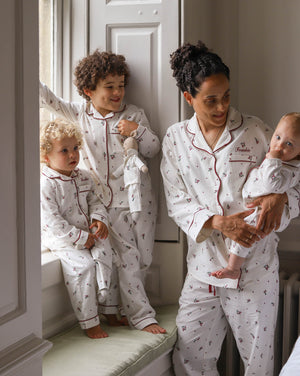 A family of four wearing matching white patterned pajamas with red piping, posed in a bright bedroom setting. The set includes an adult and three children, all wearing coordinated long-sleeve tops and pants with red trim and a small repeating motif.
