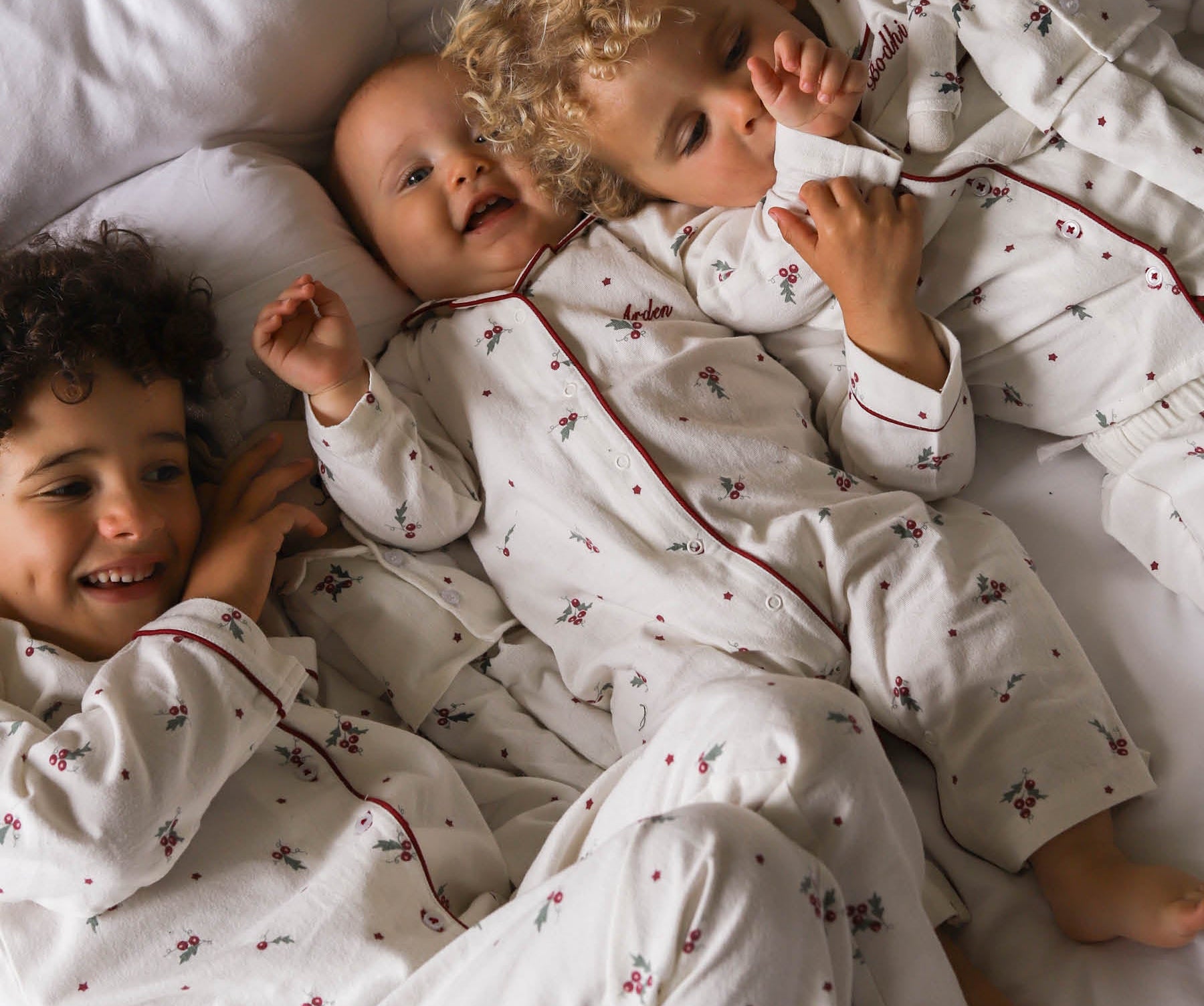 Three children lying on a bed wearing matching white long-sleeve pajamas with a small red and green floral print and dark red piping. One child is holding a white teddy bear.