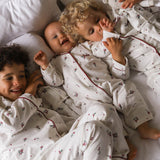 Three children lying on a bed wearing matching white long-sleeve pajamas with a small red and green floral print and dark red piping. One child is holding a white teddy bear.