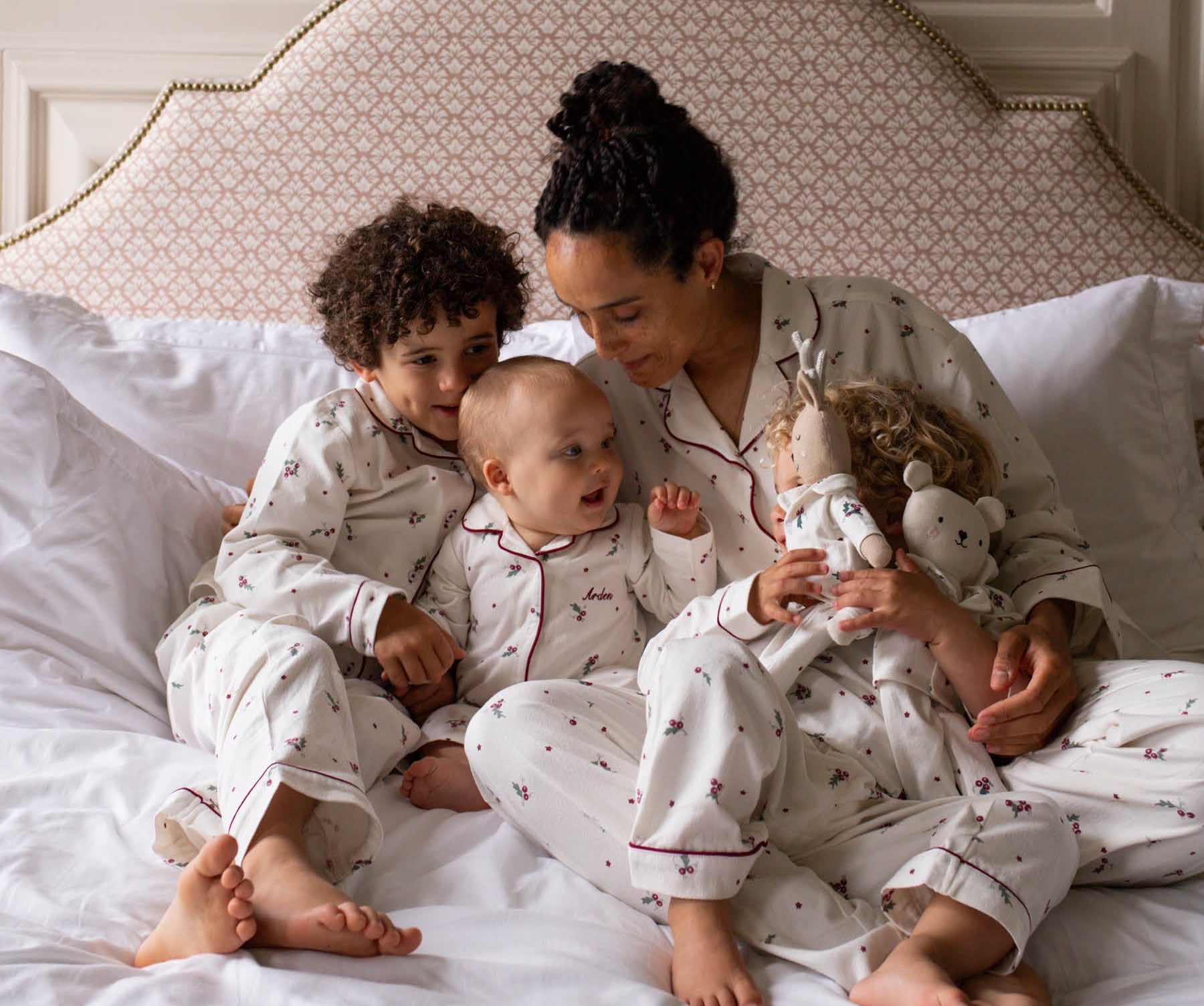 A family of four wearing matching white patterned pajamas sits together on a bed with white linens and a patterned upholstered headboard. The woman holds a doll, and the children look toward the camera.