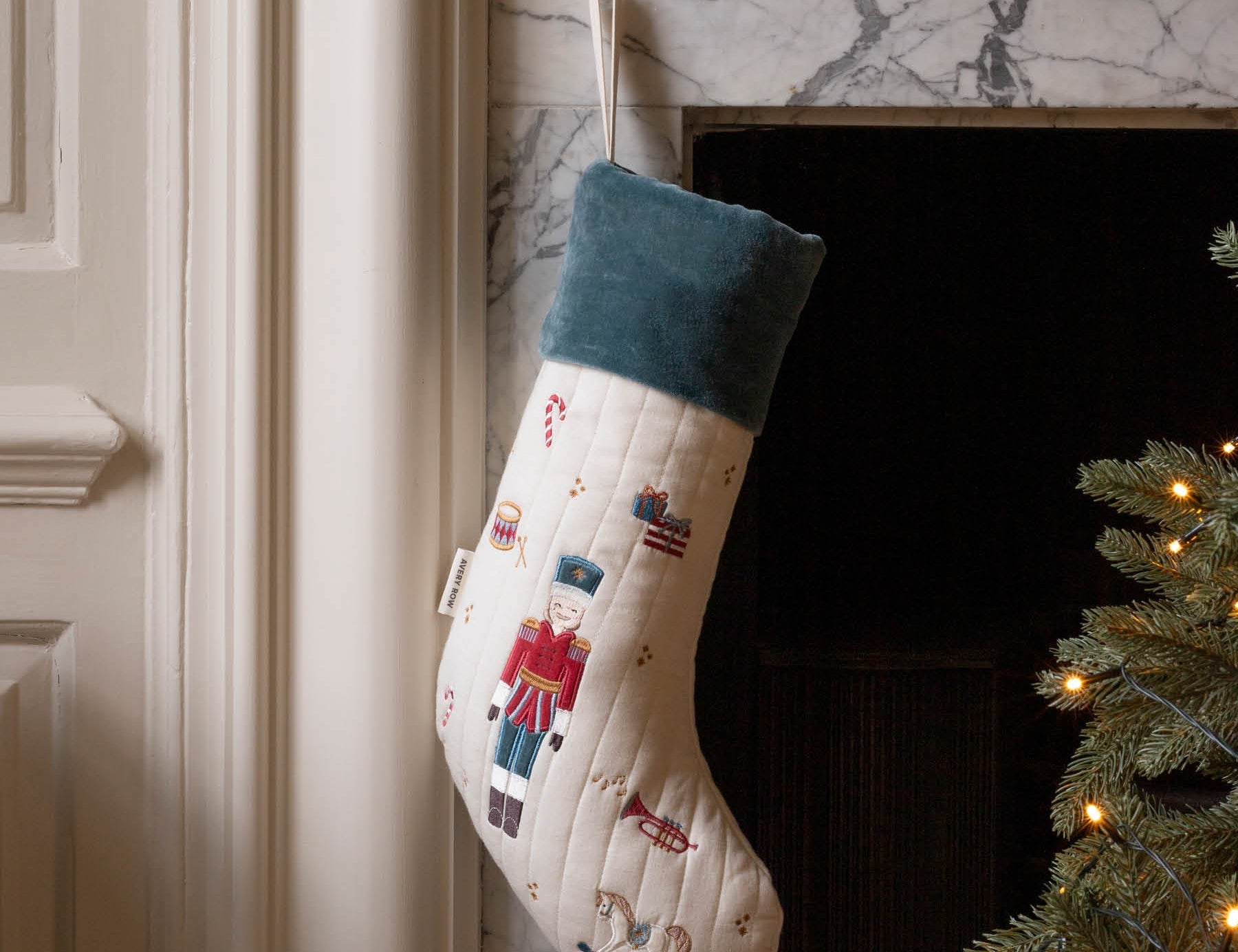 A Christmas stocking with a nutcracker design hangs in front of a marble fireplace. The stocking is cream-colored with a dark teal cuff and features embroidered nutcracker figures.