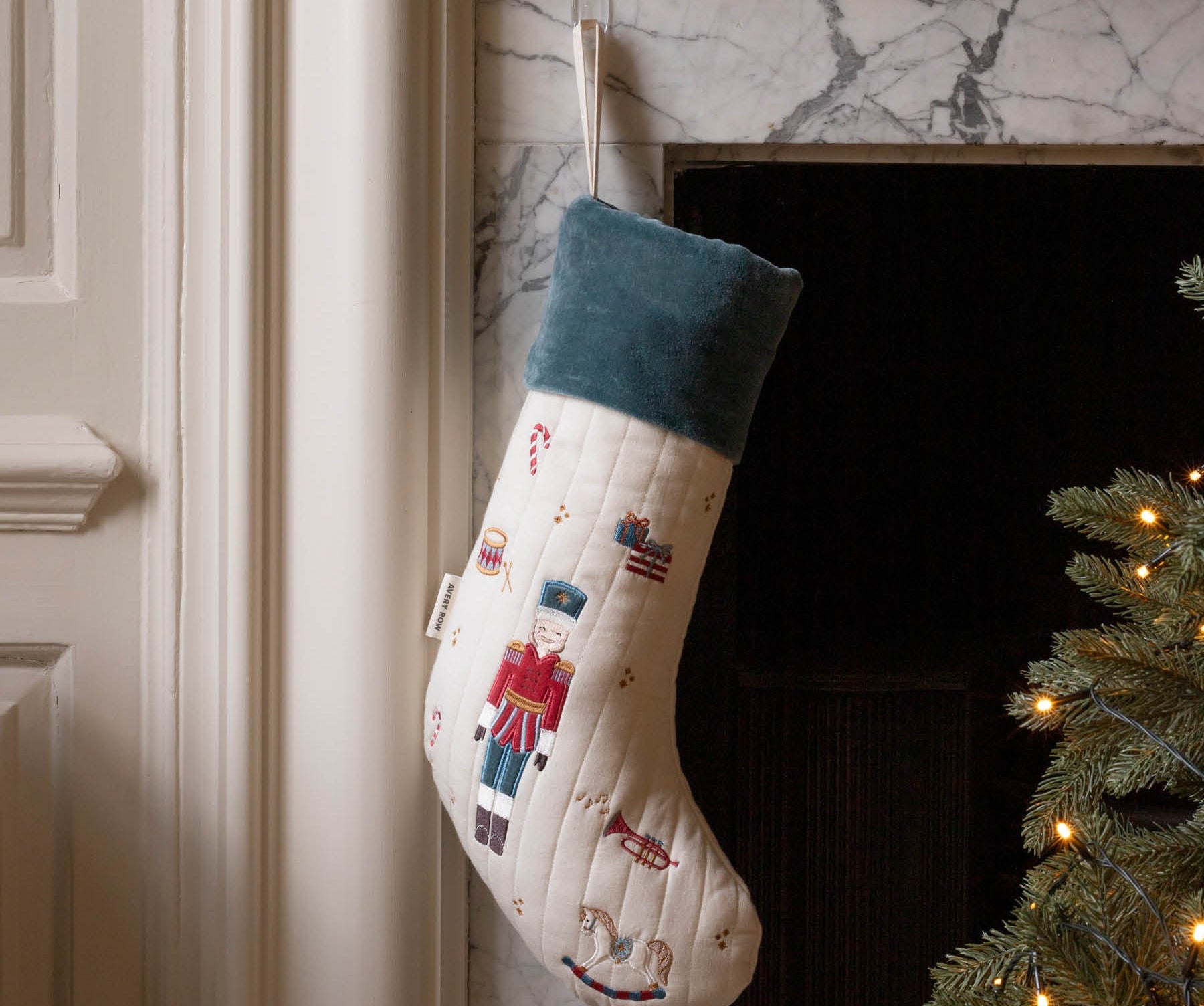 A Christmas stocking with a nutcracker design hangs in front of a marble fireplace. The stocking is cream-colored with a dark teal cuff and features embroidered nutcracker figures.