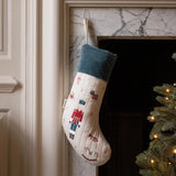 A Christmas stocking with a nutcracker design hangs in front of a marble fireplace. The stocking is cream-colored with a dark teal cuff and features embroidered nutcracker figures.