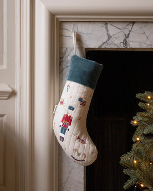 A Christmas stocking with a nutcracker design hangs in front of a marble fireplace. The stocking is cream-colored with a dark teal cuff and features embroidered nutcracker figures.