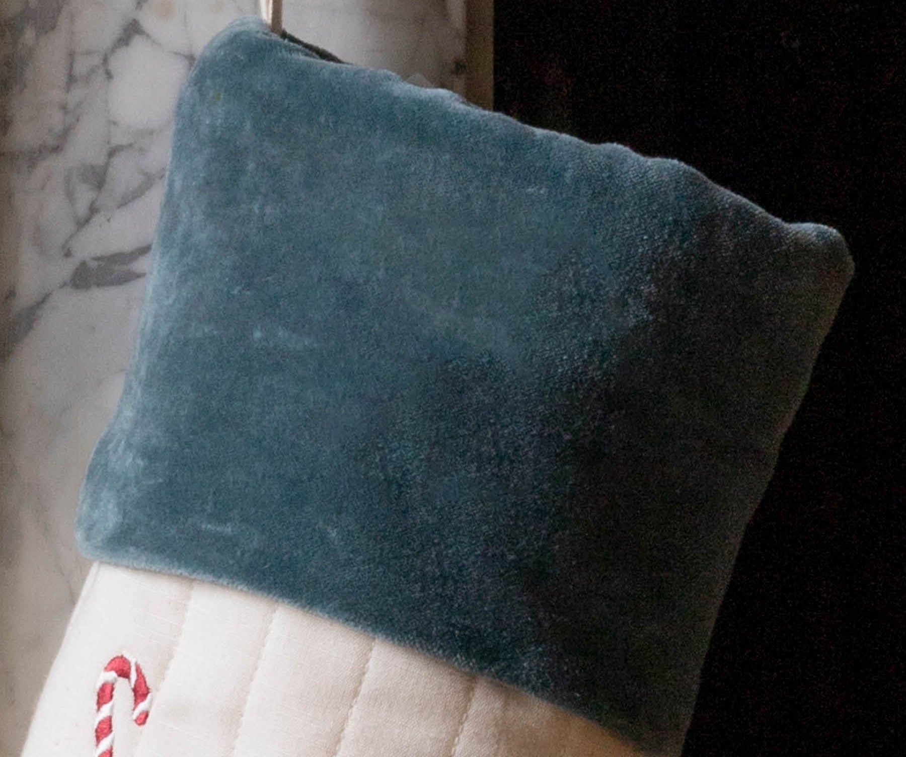 A close-up of a Christmas stocking with a dark teal velvet cuff and a cream quilted body decorated with a red-and-white candy cane motif, hanging in front of a fireplace.