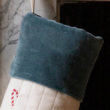 A close-up of a Christmas stocking with a dark teal velvet cuff and a cream quilted body decorated with a red-and-white candy cane motif, hanging in front of a fireplace.