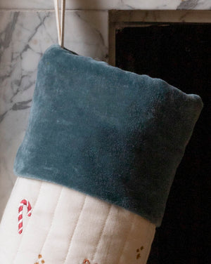 A close-up of a Christmas stocking with a dark teal velvet cuff and a cream quilted body decorated with a red-and-white candy cane motif, hanging in front of a fireplace.