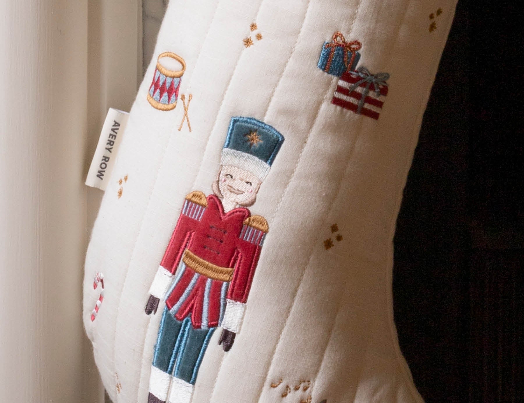 An off-white quilted Christmas stocking with embroidered festive motifs, including a central nutcracker figure in a red and blue uniform, a candy cane, a trumpet, and small gift boxes. A fabric tag on the side reads 'MERRY ROW'.