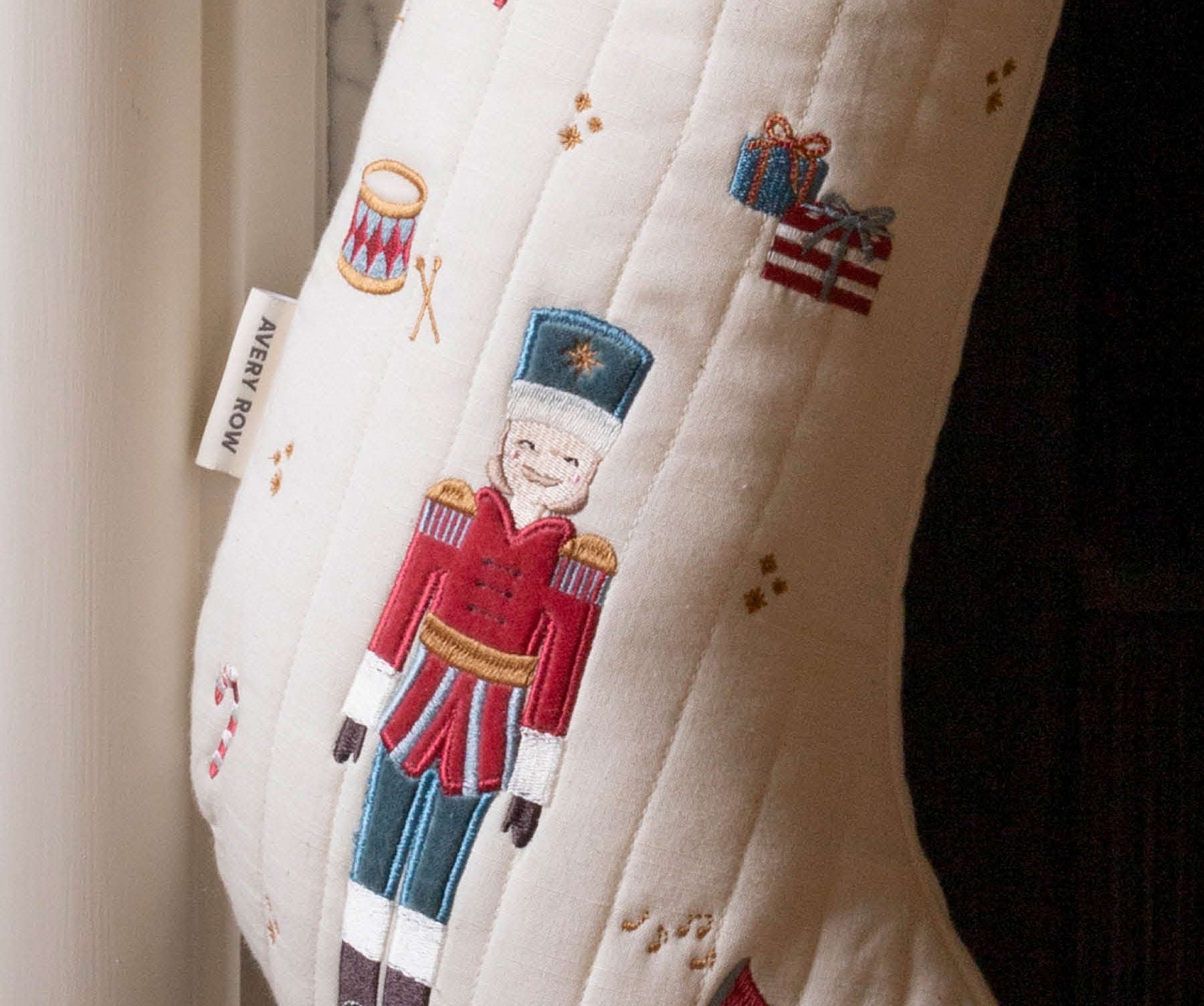An off-white quilted Christmas stocking with embroidered festive motifs, including a central nutcracker figure in a red and blue uniform, a candy cane, a trumpet, and small gift boxes. A fabric tag on the side reads 'MERRY ROW'.