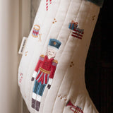 An off-white quilted Christmas stocking with embroidered festive motifs, including a central nutcracker figure in a red and blue uniform, a candy cane, a trumpet, and small gift boxes. A fabric tag on the side reads 'MERRY ROW'.
