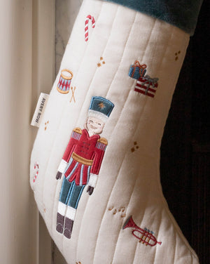 An off-white quilted Christmas stocking with embroidered festive motifs, including a central nutcracker figure in a red and blue uniform, a candy cane, a trumpet, and small gift boxes. A fabric tag on the side reads 'MERRY ROW'.