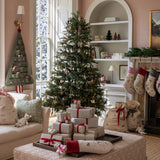 A beautifully decorated Christmas living room featuring a tall evergreen tree with lights and ornaments, wrapped gift boxes on a patterned ottoman, stockings hanging from a fireplace, and burlap Santa bags beside it.