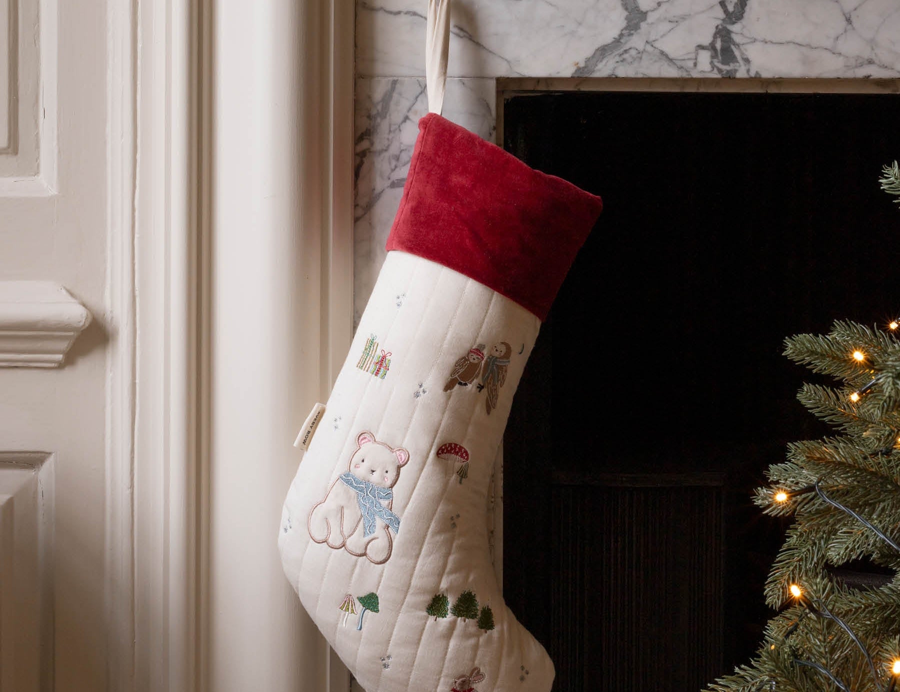 A cream-colored Christmas stocking with embroidered teddy bear motifs and a red velvet cuff, hanging in front of a marble fireplace next to a lit Christmas tree.