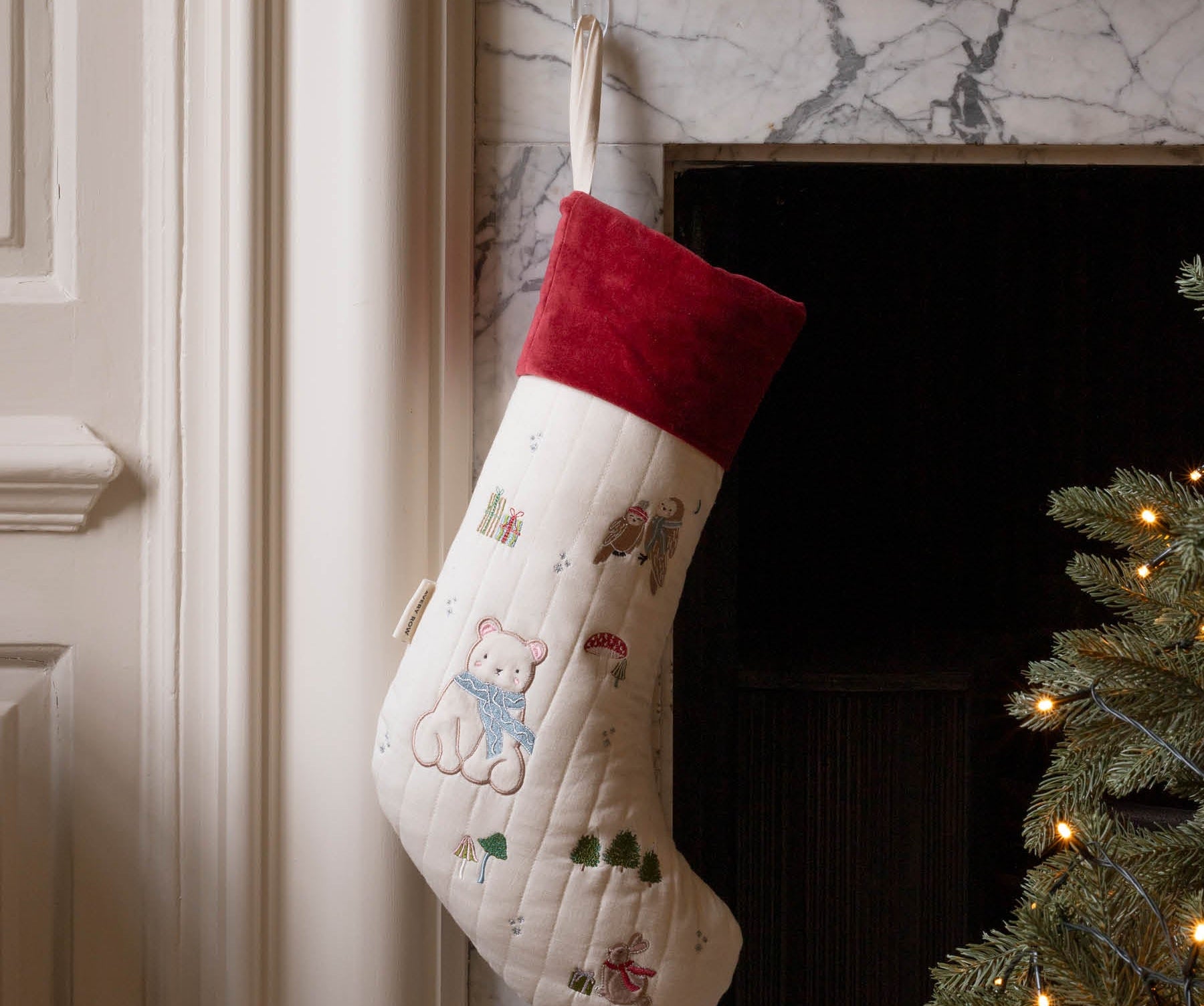 A cream-colored Christmas stocking with embroidered teddy bear motifs and a red velvet cuff, hanging in front of a marble fireplace next to a lit Christmas tree.